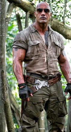 the rock is standing in front of some trees and wearing gloves with his hands on his hips