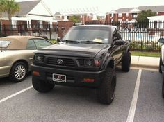 a black truck parked in a parking lot next to other cars and buildings with palm trees behind it