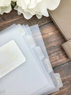 three clear folders with gold foil lettering on them and a white flower in the background