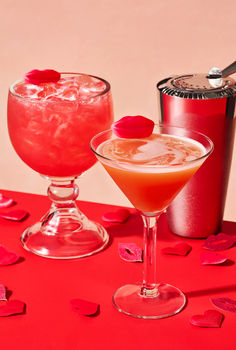 two cocktails on a red table with confetti and hearts scattered around them