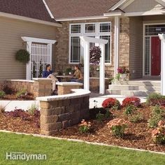 two people sitting on the front porch of a house with landscaping around them and flowers