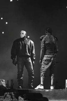 two men standing on top of a stage with lights in the sky behind them and one man wearing a mask