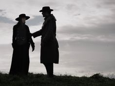 two men standing on top of a grass covered hill next to each other wearing long coats and hats