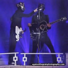 two men in black cats costumes are playing guitar and singing on stage with blue lights behind them