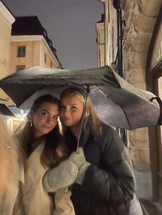 two young women standing under an umbrella in the rain at night, one holding her arm around the other's shoulder