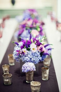 a long table with purple and white flowers in vases next to shot glasses on it