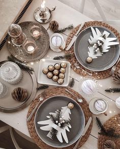 the table is set for christmas dinner with silver and white plates, pineconi