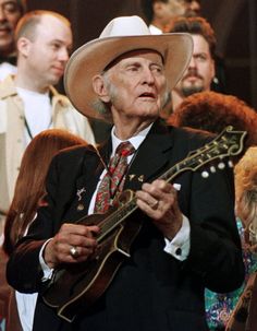 an old man wearing a cowboy hat and holding a guitar in front of a group of people