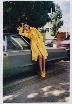 a woman leaning on the hood of a car wearing a yellow trench coat and heels