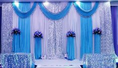 a wedding stage decorated with blue and white drapes, purple flower arrangements, and silver sequins