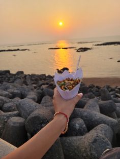 a person holding food in their hand on the beach at sunset or sunrise with seagulls flying overhead