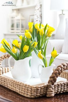 yellow tulips are in white pitchers on a wicker tray