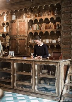 a woman sitting at a counter in a room with many shelves and jars on the wall