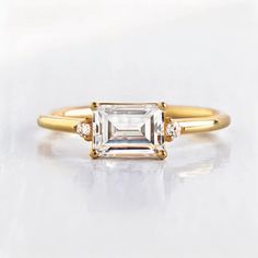 a gold ring with an emerald cut diamond and three diamonds on the band, sitting on a white surface