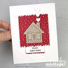 a hand holding up a christmas card with a house on it and a heart hanging from the top