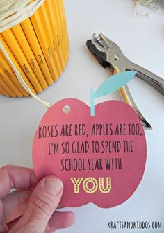 a hand holding a pink apple shaped paper tag with writing on it and some pencils in the background