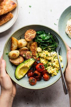 two hands holding a bowl of food with eggs, potatoes and spinach on the side