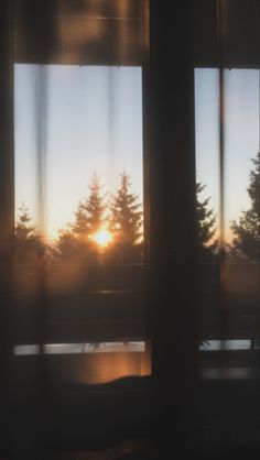 the sun is setting through some curtains in front of a window with pine trees outside