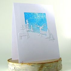 a card with reindeers and trees on it sitting on top of a piece of wood