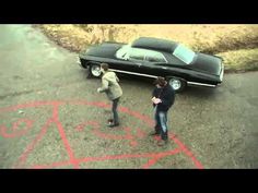 two men standing in front of a black car with red graffiti on the ground next to it