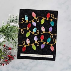 a card with christmas lights on it next to some pine cones and evergreen branches in the background