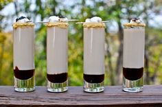 four glasses filled with chocolate pudding and marshmallows on a wooden table in front of trees