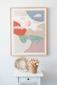 a small white table with a basket and flowers on it next to a wall hanging