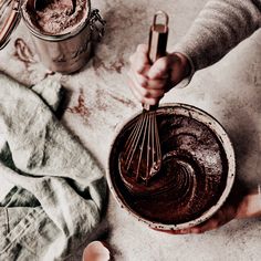 a person holding a whisk over a bowl filled with chocolate cake batter on top of a table