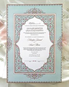 an ornate wedding card with pink and blue trimmings on the edges, in front of white satin