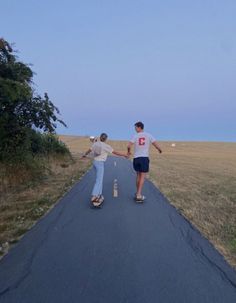 two people skateboarding down a road in the middle of an open field with trees