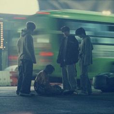 three boys are standing on the side of the road near a bus and another boy is laying on the ground