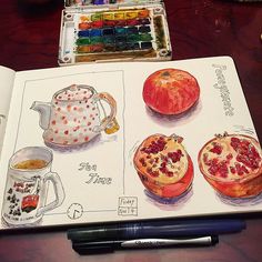 an open book with watercolors on it next to a cup of coffee and two pomegranates