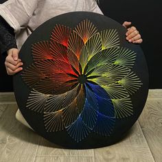 a person holding up a circular artwork made out of colored sticks and wire on top of a wooden table