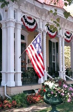 an american flag hanging on the front porch of a house with flowers and potted plants