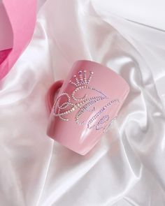 a pink mug with a tiara on it sitting on a white satin covered surface