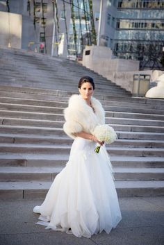 a woman in a white dress and fur stole holding a bouquet of flowers on some steps