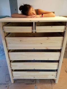 a woman laying her head on top of a wooden cabinet with drawers and shelves underneath it