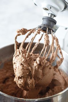 a metal bowl filled with chocolate frosting on top of a white countertop next to a mixer