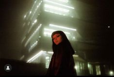 a woman standing in front of a tall building with lights shining down on her face