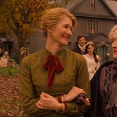 two women standing next to each other in front of a house with people around them