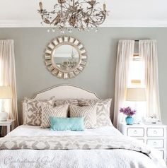 a white bed sitting under a chandelier in a bedroom next to two windows