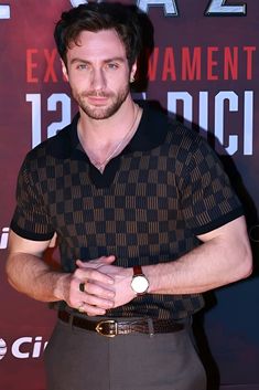 a man standing in front of a red wall wearing a black shirt and brown pants