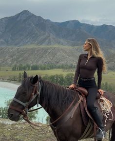 a woman riding on the back of a brown horse next to a lake and mountains