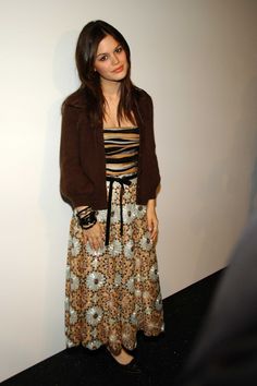 a woman standing in front of a white wall wearing a brown cardigan and floral dress