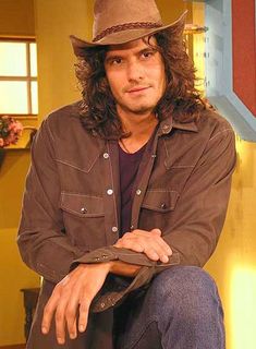 a man with long hair wearing a brown hat and jacket sitting in front of a sign