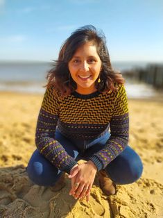 a woman sitting on top of a sandy beach next to the ocean with her hands in her pockets