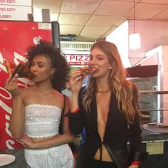 two women standing in front of a pizza stand eating donuts and drinking sodas