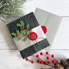 two christmas cards with holly leaves and red buttons on them, next to pine cones