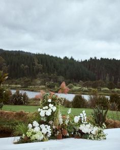 flowers and greenery are arranged on a table outside near a lake in the background