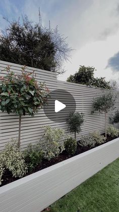 a white fence with some trees and bushes in front of it on the side of a building